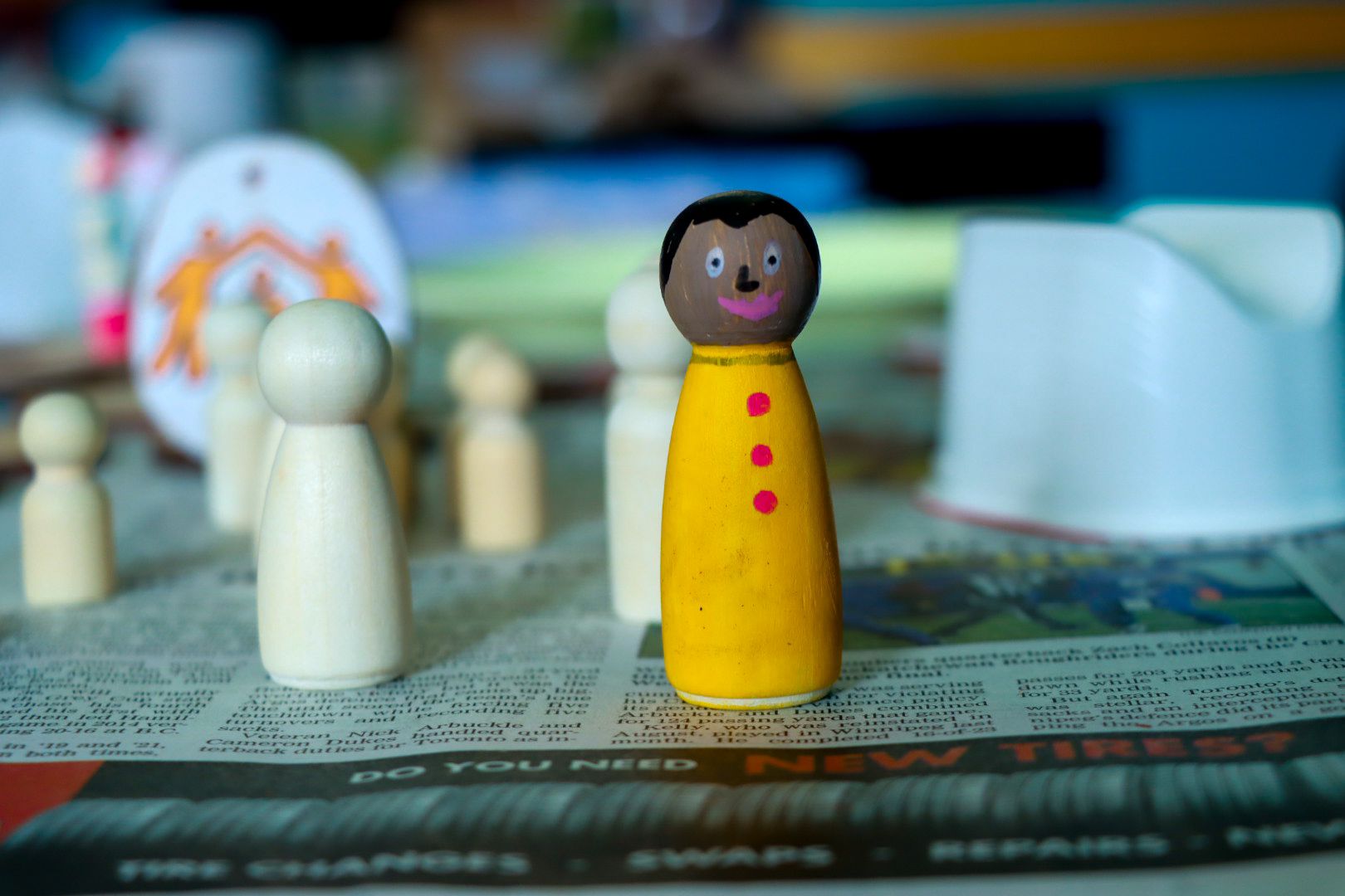 Painted wooden figure with a smiling face and yellow outfit among unpainted wooden figures on a table.
