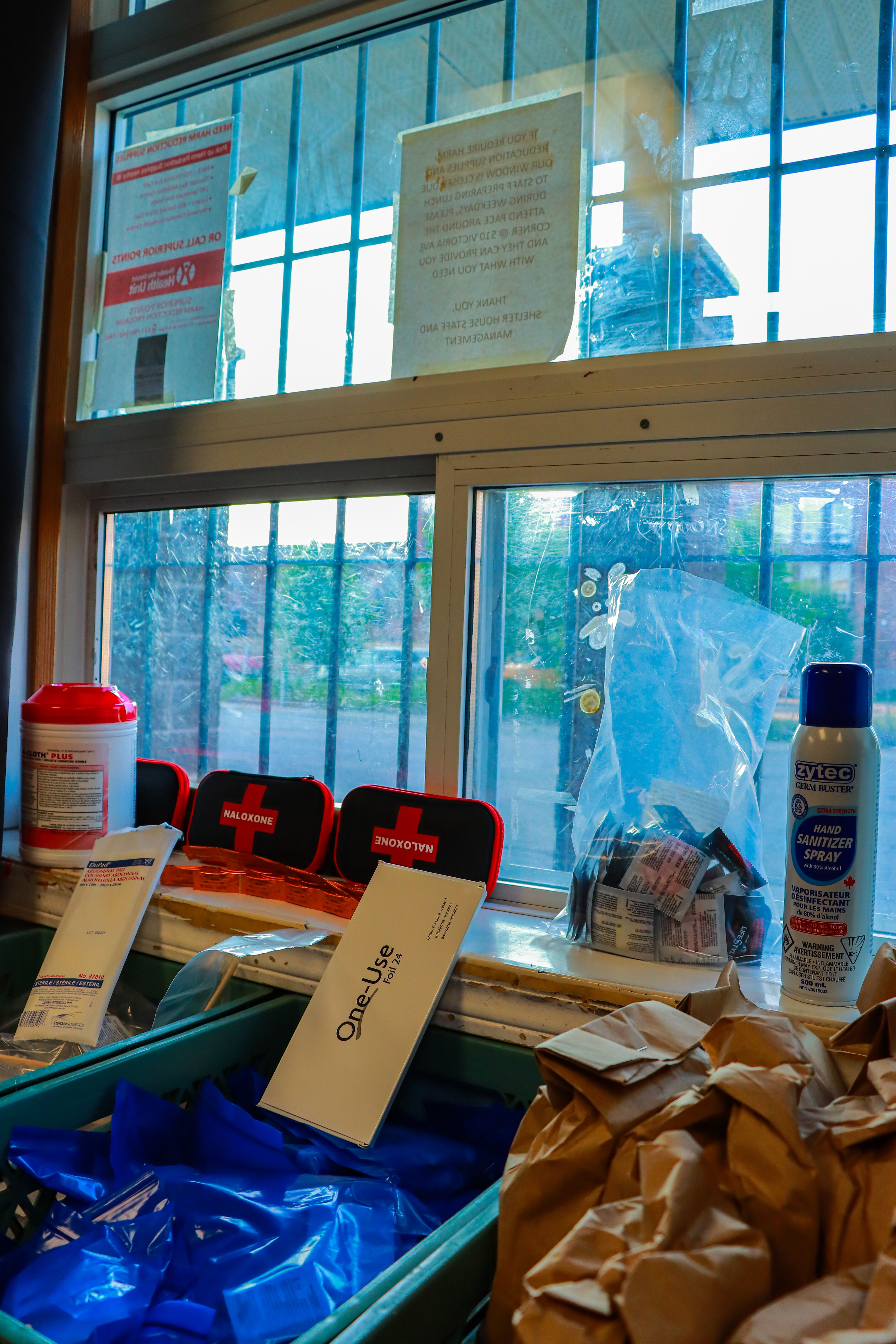 Harm reduction supplies on a windowsill, including naloxone kits, sanitizer spray, gloves, and paper bags.