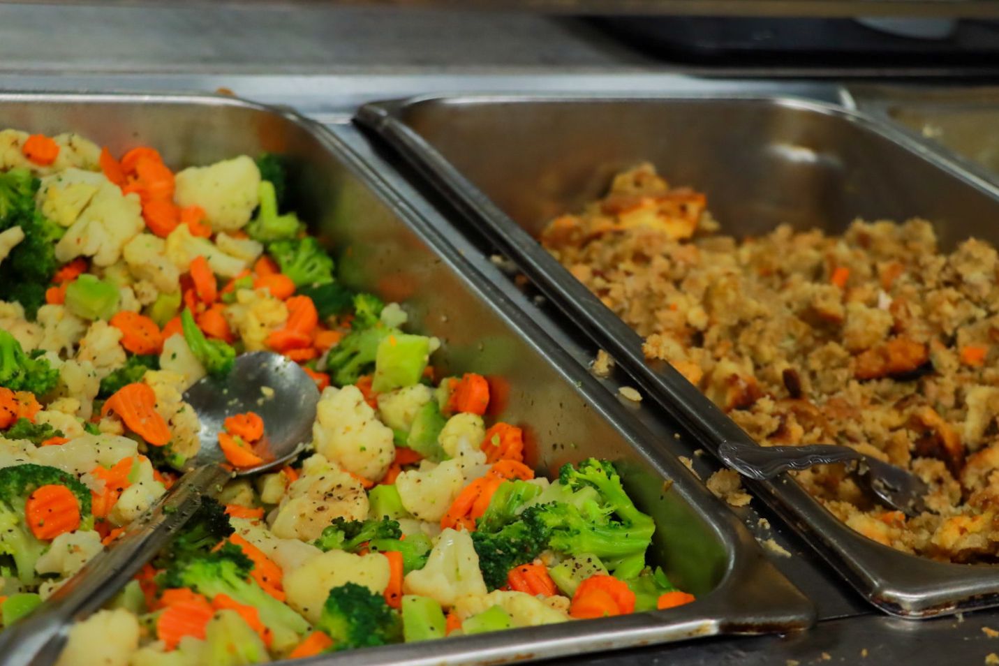 Serving trays filled with cooked mixed vegetables and stuffing in a kitchen setting.