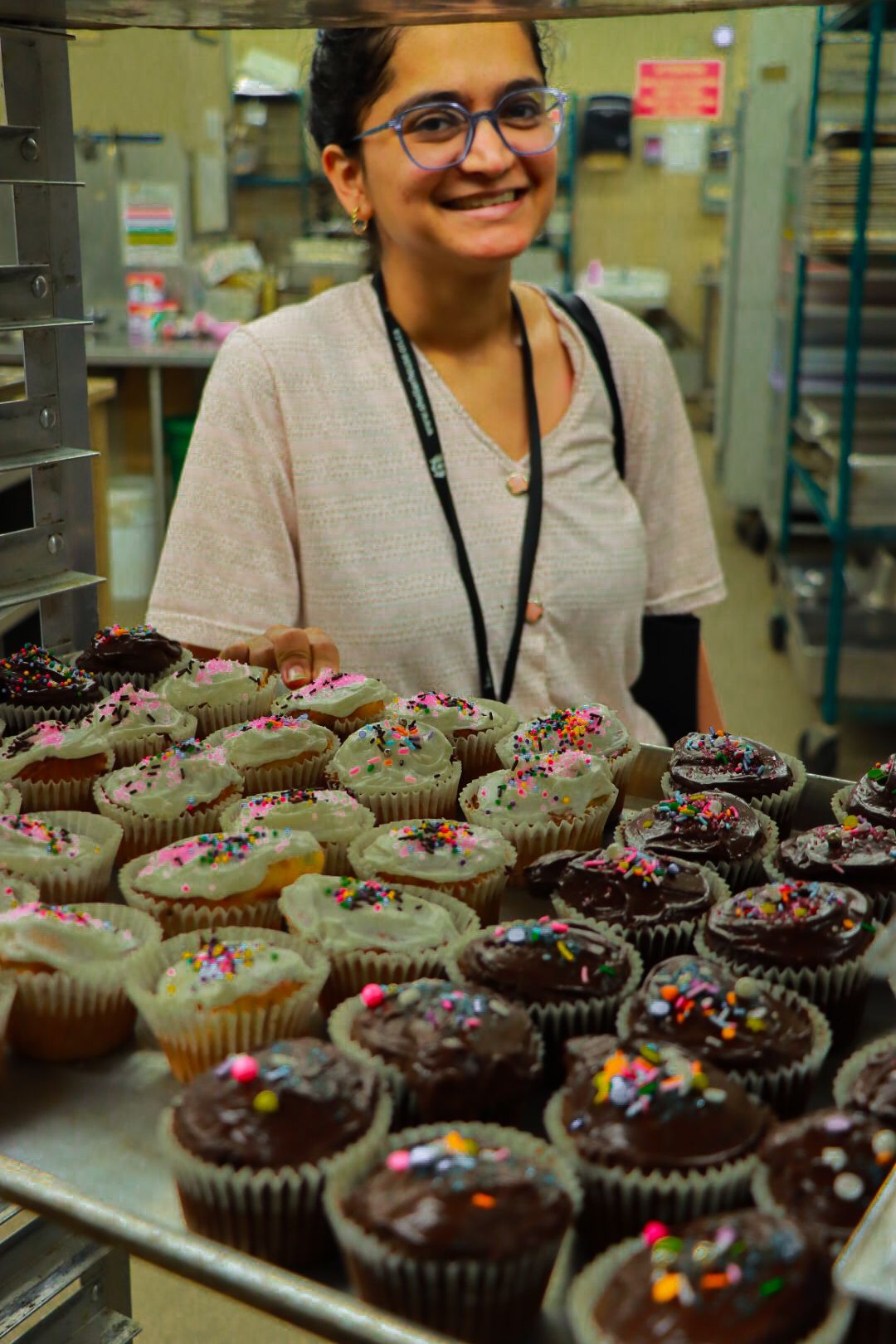 Smiling person in a kitchen holding a tray of cupcakes decorated with sprinkles.