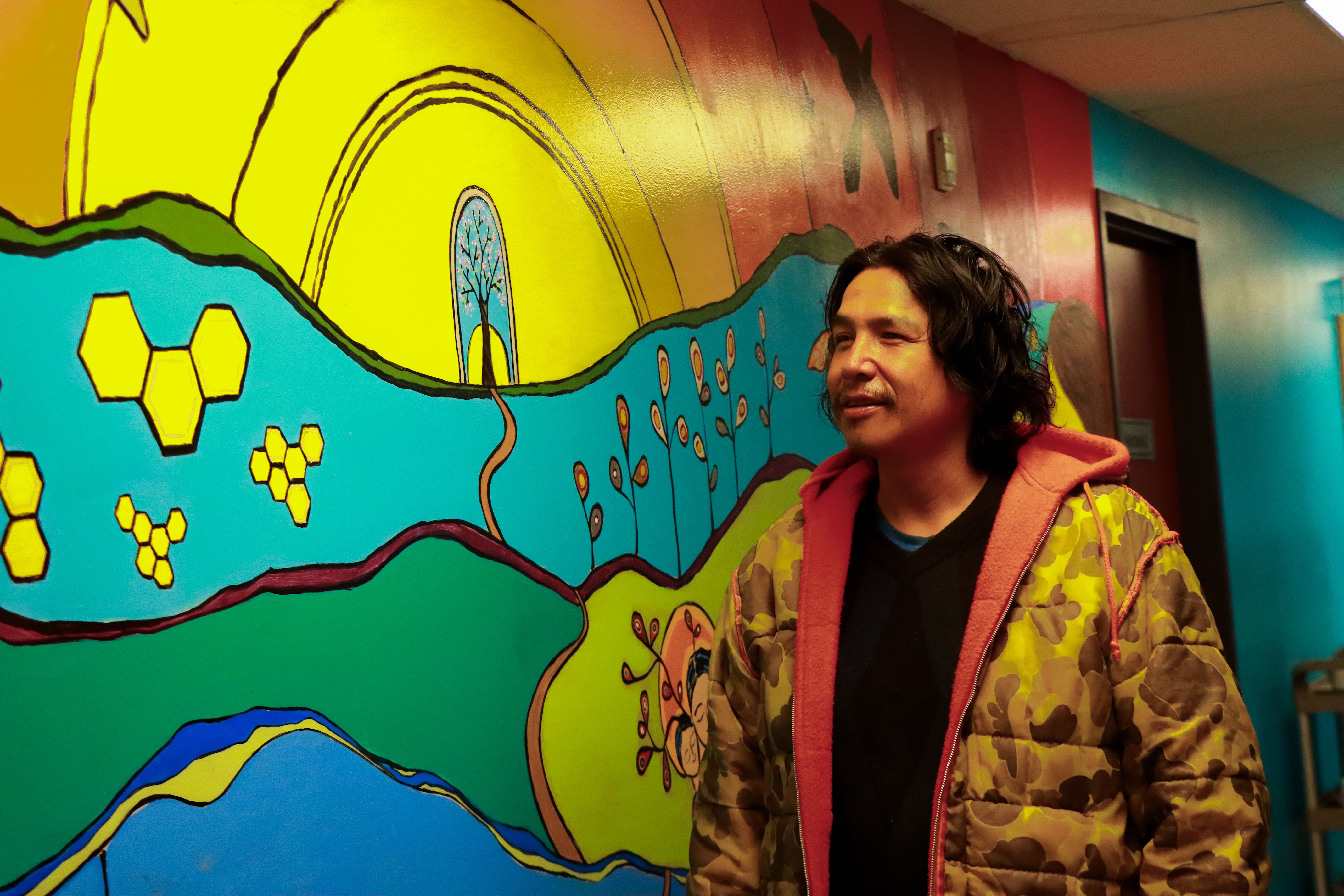 Man in a camouflage jacket standing in front of a colorful mural with bright yellow, blue, and green designs.