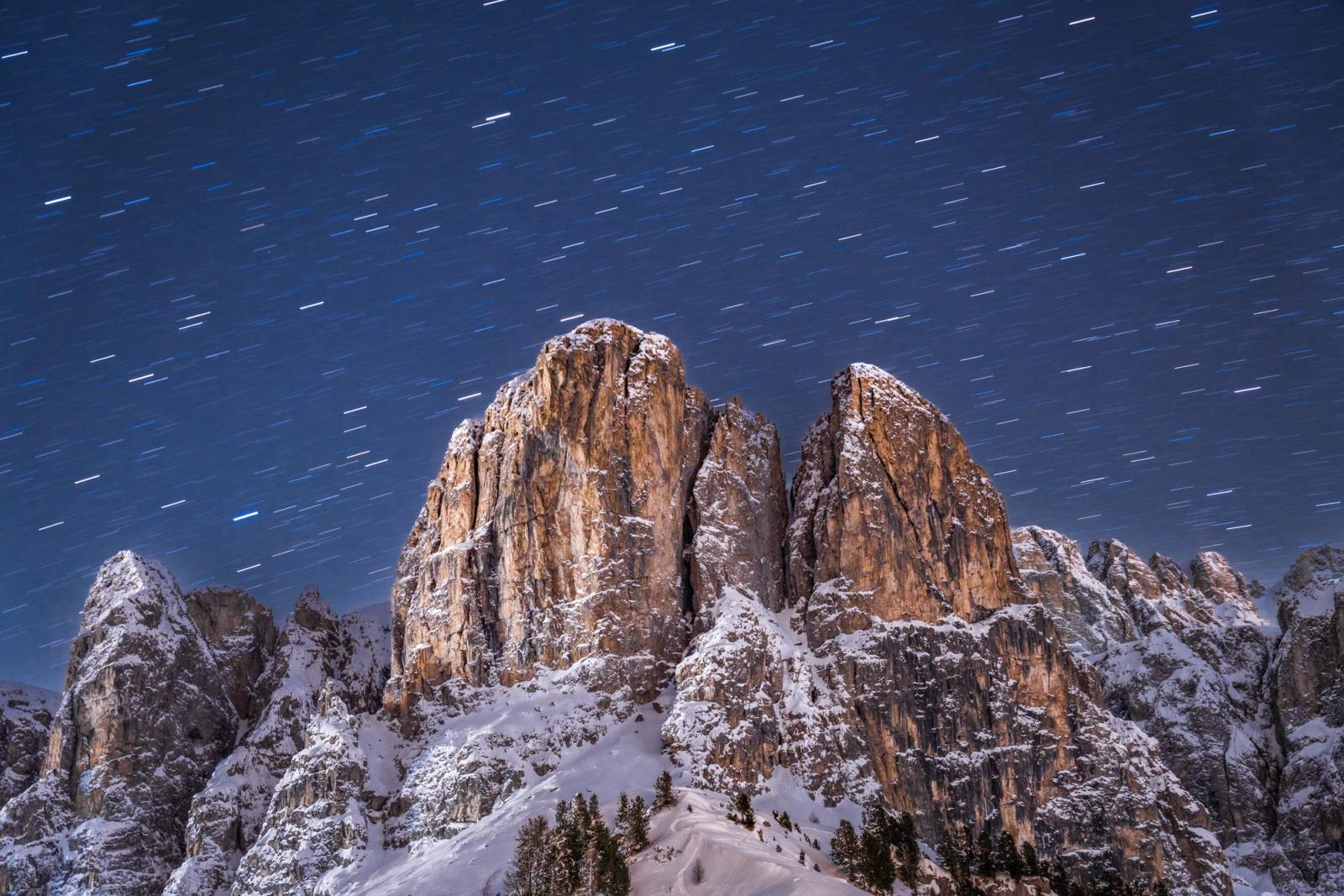 Star trails arc across a deep blue winter sky above the illuminated limestone towers of the Dolomites, their warm amber faces contrasting with snow-laden slopes below. A long exposure of several minutes has resolved individual stellar paths into short parallel dashes, the moon providing the ambient light that reveals every ledge and crack in the rock face. Conifers at the snowline anchor the composition and restore a sense of scale.