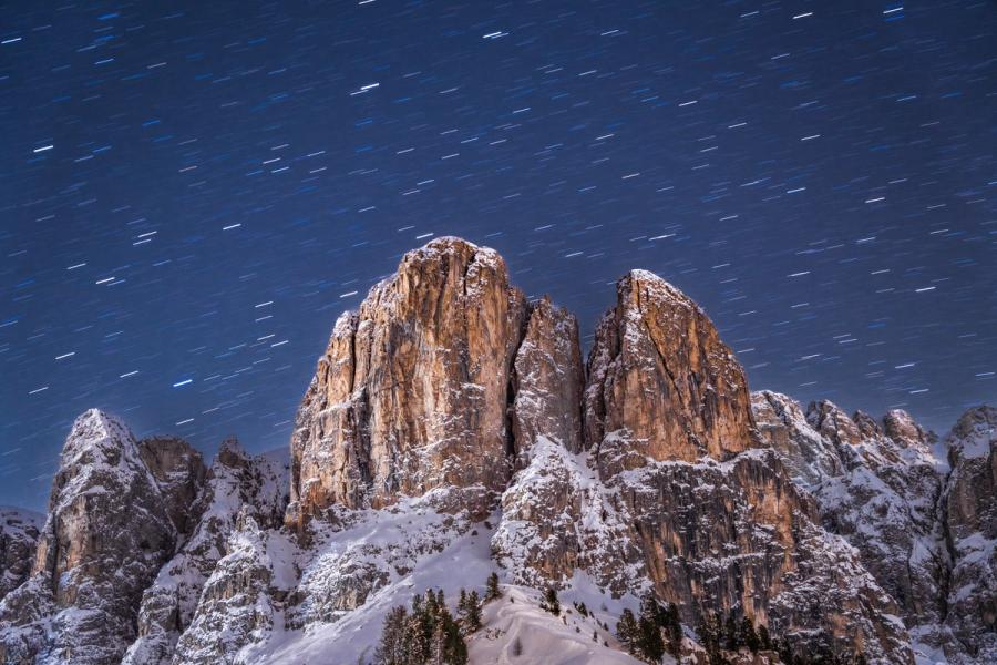 Star trails arc across a deep blue winter sky above the illuminated limestone towers of the Dolomites, their warm amber faces contrasting with snow-laden slopes below. A long exposure of several minutes has resolved individual stellar paths into short parallel dashes, the moon providing the ambient light that reveals every ledge and crack in the rock face. Conifers at the snowline anchor the composition and restore a sense of scale.