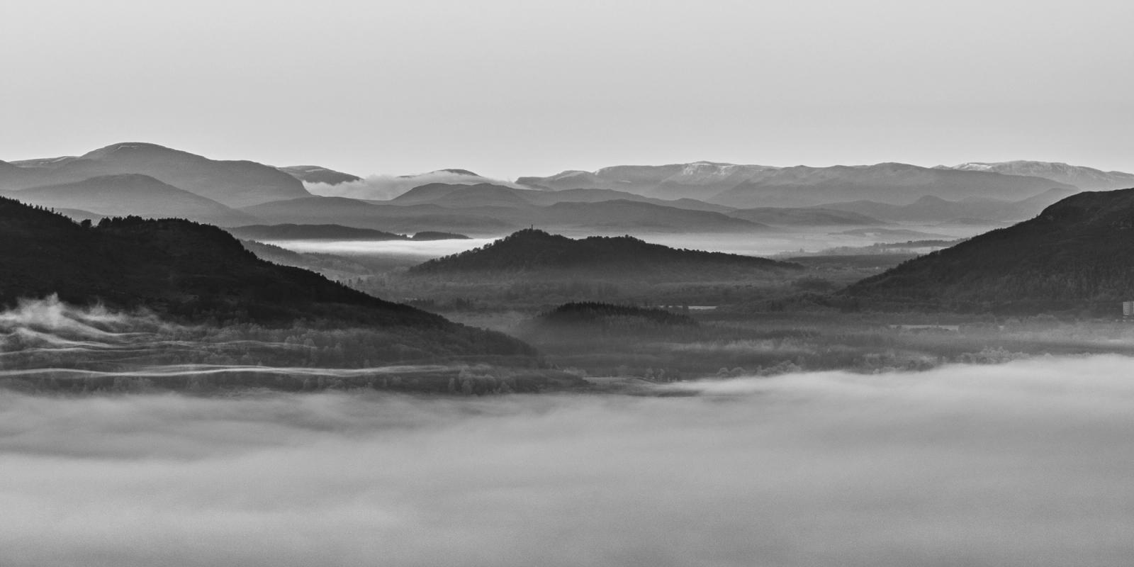 A temperature inversion fills the Spey Valley on Christmas morning, submerging the low ground in dense fog while successive ridgelines recede into haze above Boat of Garten. The monochrome conversion distils the scene to pure tonal recession, with a hilltop structure â possibly a monument â emerging as the sole punctuation in the middle distance. Aerial elevation reveals the full depth of the layering in a way unavailable from the valley floor.