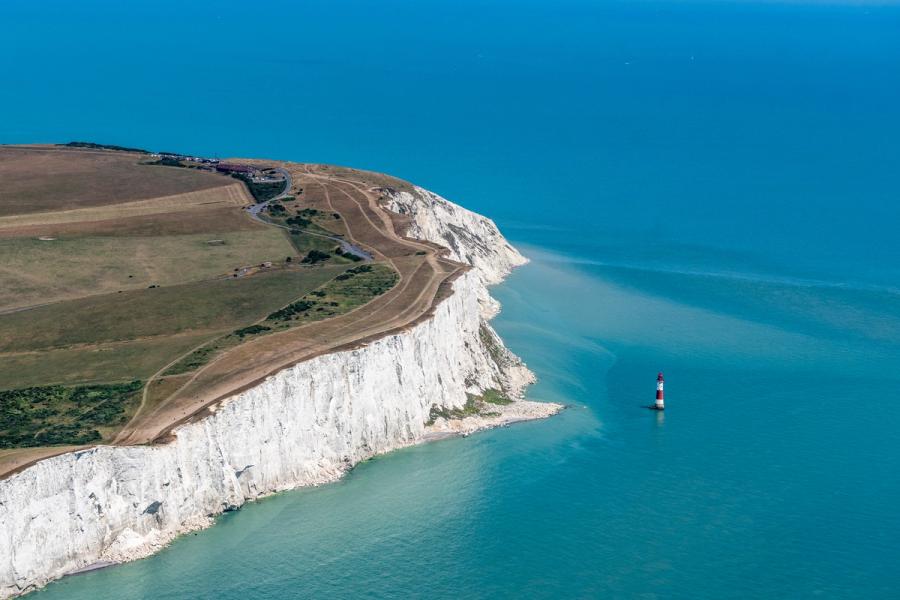 Beachy Head lighthouse stands isolated at the base of the chalk cliffs, dwarfed by the headland's sweeping face. Shot from a high lateral vantage, the image pits the geometry of eroded chalk strata against the flat blue of the English Channel. The midday light is clean but harsh, flattening shadow detail across the cliff face.