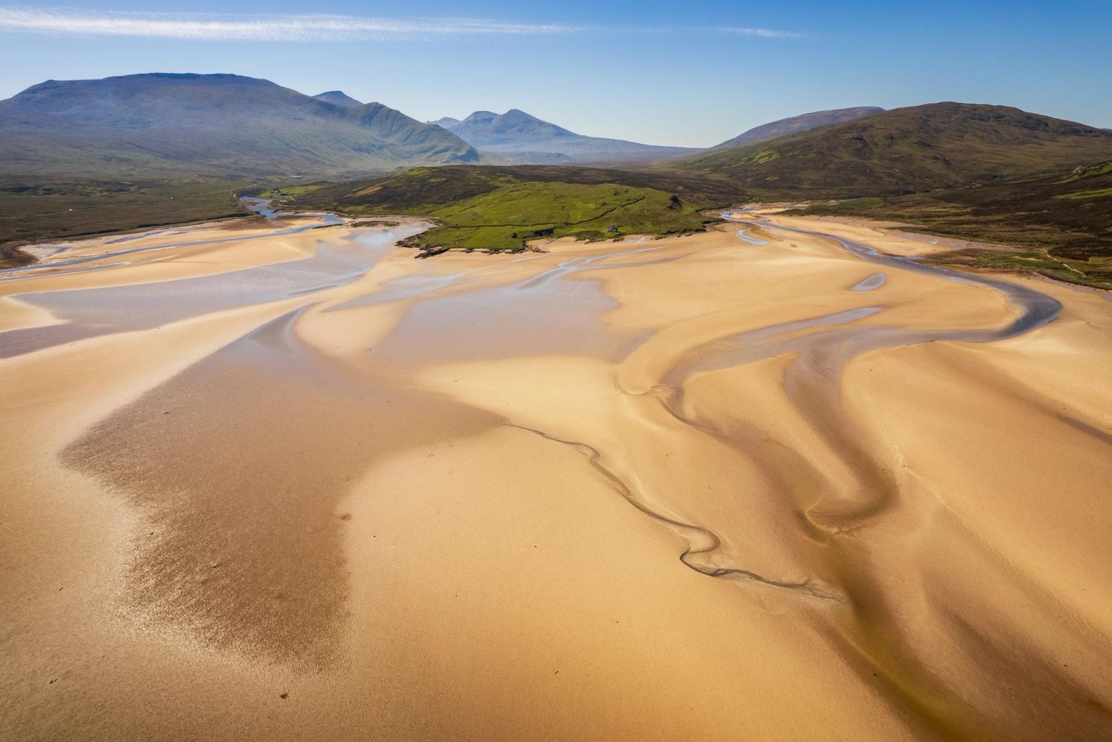 At low tide, braided channels carve sinuous lines across the tidal flats of a sea loch in Durness, Sutherland, the warm golden sand contrasting with thin slivers of pewter-grey water. Beyond, a patchwork of moorland and machair gives way to a receding line of mountains under a clear spring sky. The aerial vantage flattens the scene into near-abstraction while retaining the scale of the Highland interior.