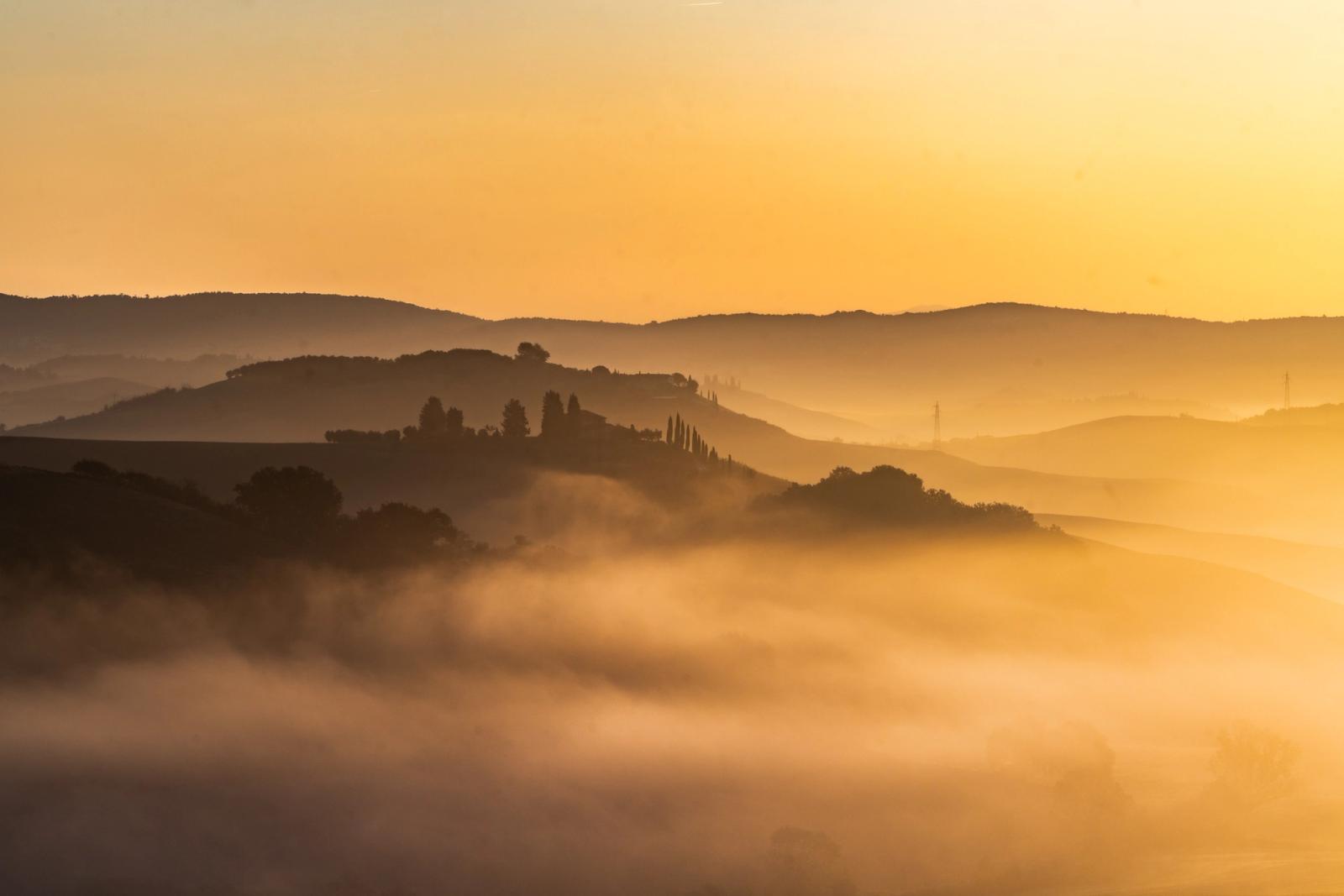 Layers of ground fog pool between rolling hills at first light, the warm amber sky backlighting successive ridgelines into graduated silhouettes. Cypress trees on a central hill anchor the composition against the dissolving mist below. The scene has the character of the Tuscan or central Italian Apennine landscape at autumn dawn.