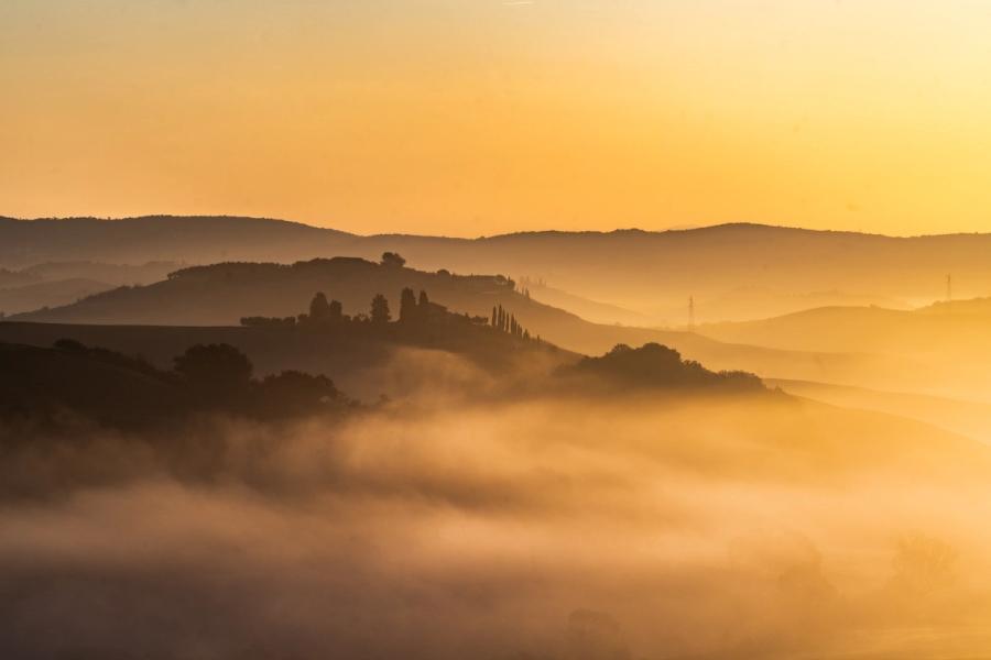 Layers of ground fog pool between rolling hills at first light, the warm amber sky backlighting successive ridgelines into graduated silhouettes. Cypress trees on a central hill anchor the composition against the dissolving mist below. The scene has the character of the Tuscan or central Italian Apennine landscape at autumn dawn.