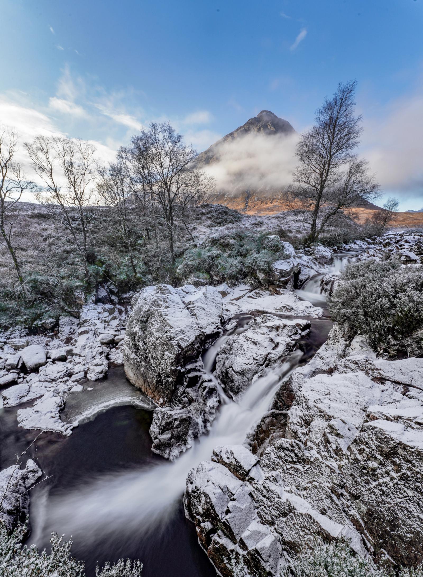 The River Coupall cascades over snow-dusted boulders below Buachaille Etive MÃ²r, its peak half-consumed by cloud. A long exposure renders the water as silk against the stillness of the frosted rockscape, the bare birches framing the pyramid summit with quiet insistence.
