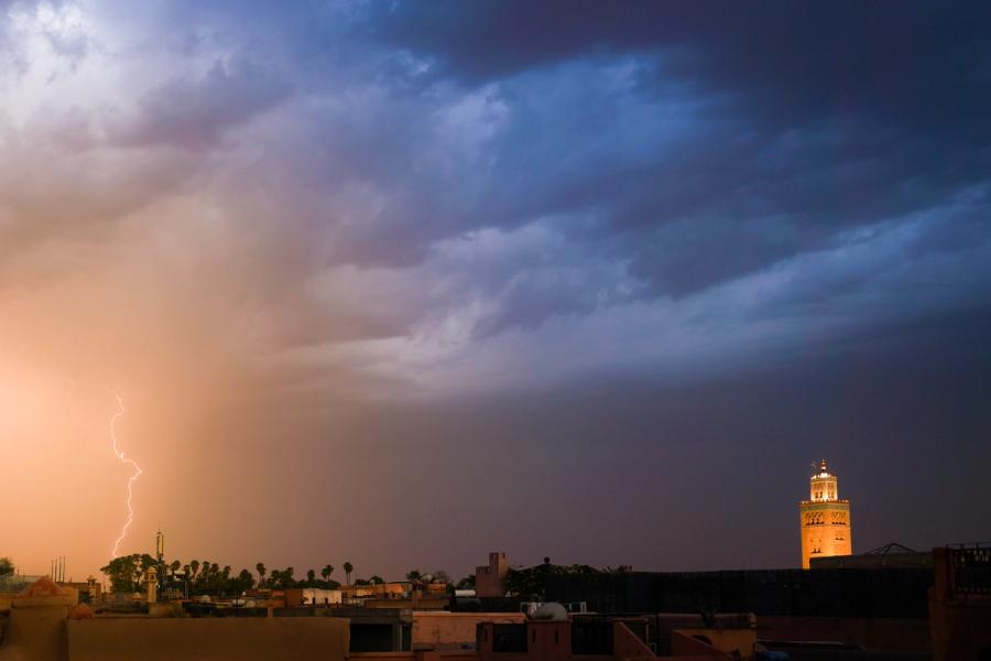A forked lightning bolt strikes beyond the rooftops as a summer storm rolls across a North African medina at dusk. The illuminated minaret of the Koutoubia Mosque anchors the right frame against a bruised, layered sky, while warm sodium light from the city below stains the rain-curtain amber to the left. The tension between the artificial warmth and the cold storm front gives the image an unusual chromatic and dramatic coherence.