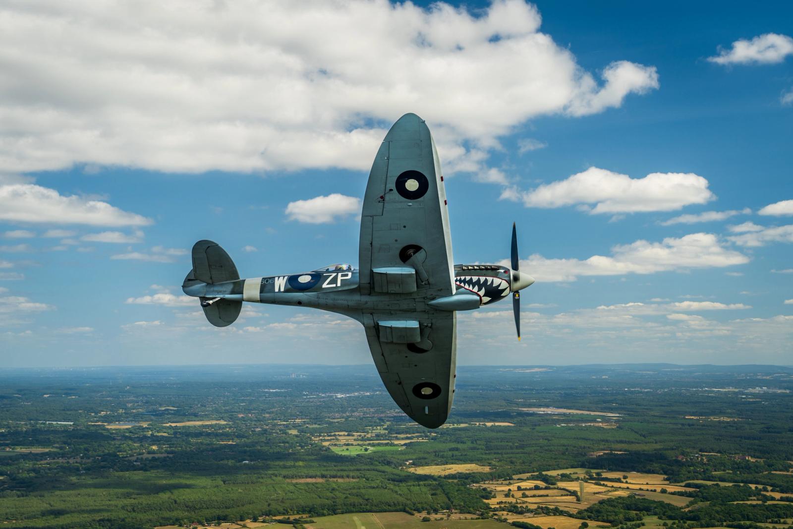 A Supermarine Spitfire in RAF markings â codes ZP-W and a shark-mouth nose detail â banks steeply above a patchwork of summer fields and woodland. The air-to-air perspective places the aircraft centre-frame against a broken cumulus sky, the rolling English lowlands receding to a distant horizon beneath it.