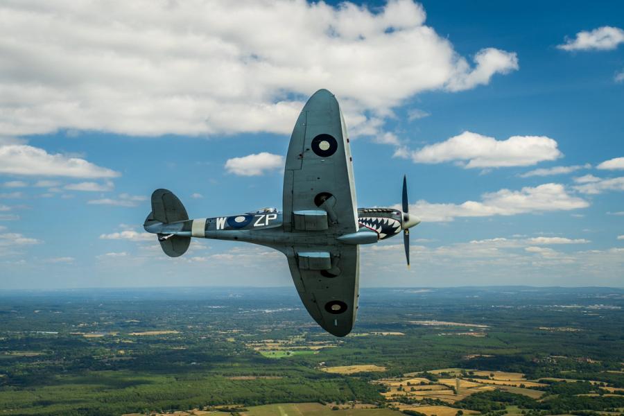 A Supermarine Spitfire in RAF markings â codes ZP-W and a shark-mouth nose detail â banks steeply above a patchwork of summer fields and woodland. The air-to-air perspective places the aircraft centre-frame against a broken cumulus sky, the rolling English lowlands receding to a distant horizon beneath it.