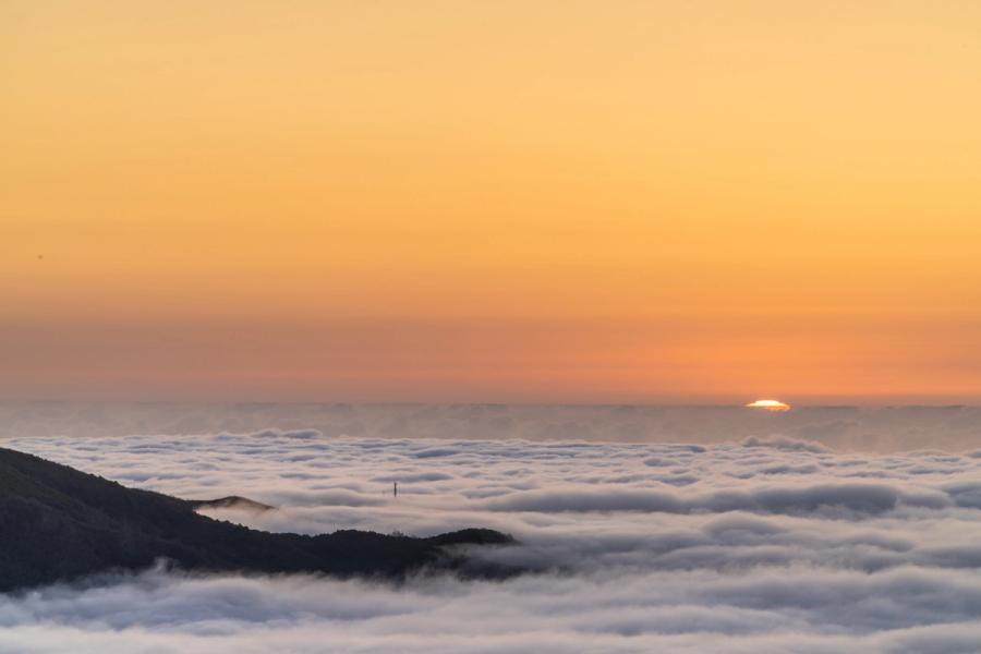 A temperature inversion buries the valleys beneath a level sea of cloud as the sun descends to the horizon, its disc just visible above the fog layer. Mountain ridges break through the white surface in the foreground, anchoring the scene. The unbroken amber sky above gives the image a clean, restrained luminosity.