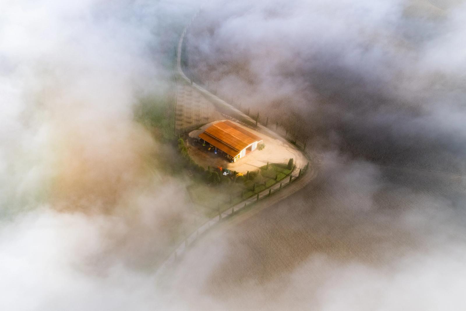 A farmstead near Monteroni d'Arbia emerges through a break in low-lying fog, its terracotta roof caught in early morning light while the surrounding Tuscan fields dissolve into cloud. The circular driveway and fence line anchor the composition, framing a moment of fleeting clarity within the inversion layer.
