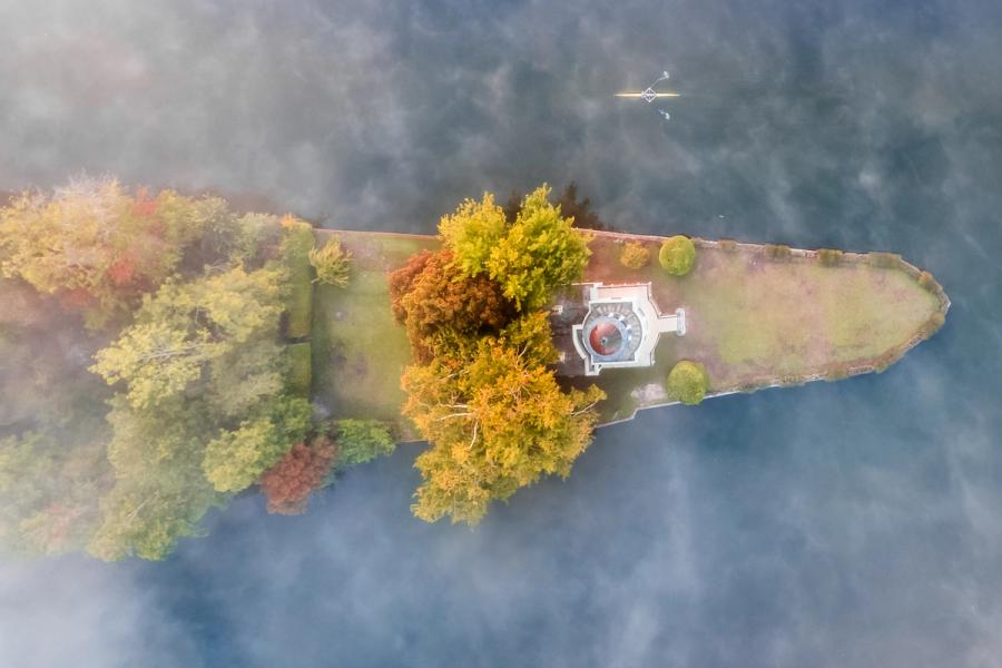A small ornamental island on the Thames near Henley, viewed directly from above through drifting autumn mist. The neoclassical pavilion sits at the island's centre, surrounded by trees in full turn, while a lone sculler passes on the still water beyond. The low cloud partially obscuring the frame gives the image an accidental painterly quality.