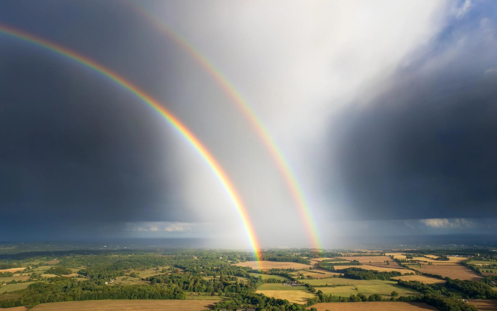 Two simultaneous rainbows converge toward the horizon above the patchwork farmland and woodland of the Thames Valley, photographed from altitude during the passage of a summer storm. The compressed aerial perspective renders both arcs as structural elements against a dark cumulonimbus backdrop, with an unusual shaft of bright sky trapped between them. Captured near Henley-on-Thames in late afternoon light, September 2025.