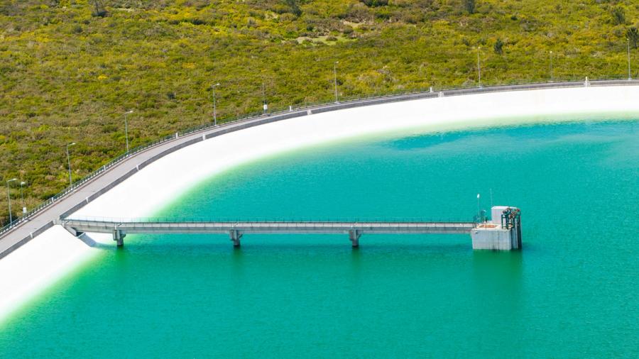 The curved face of the Calheta reservoir dam sweeps across the frame from an aerial vantage, its white concrete wall dividing vivid turquoise water from the scrubland hillside above. A narrow access bridge extends perpendicular to the intake tower, anchoring the geometric composition. Shot in bright overcast light that suppresses shadow and renders the water colour with unusual clarity.