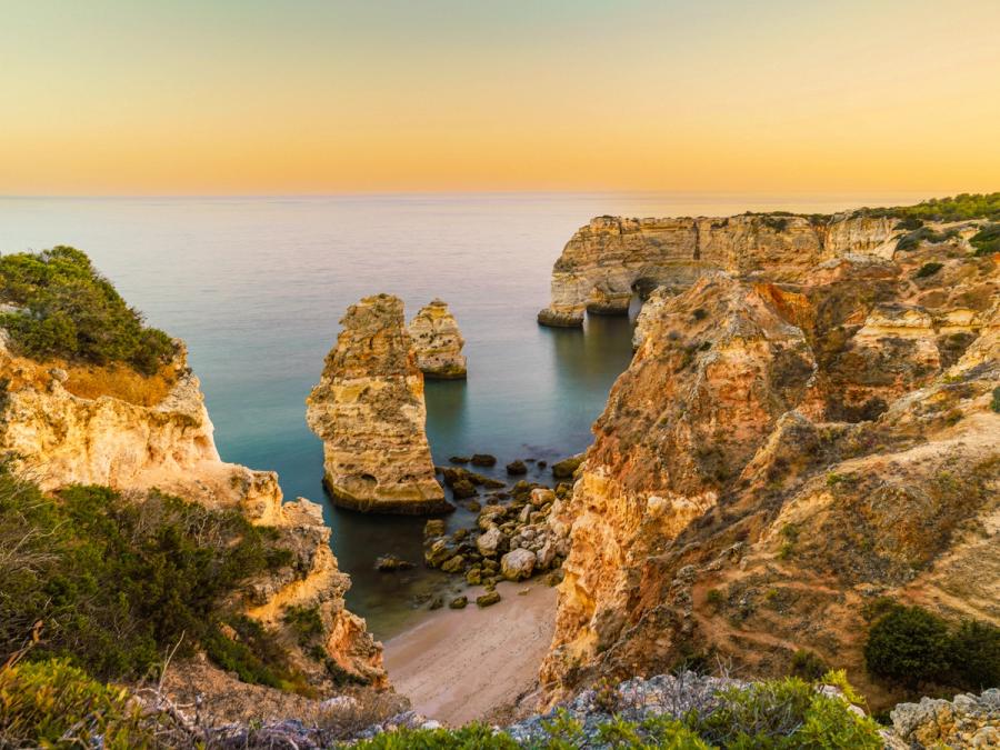 Twin sandstone stacks rise from a sheltered cove as the last light drains from a cloudless sky. The long exposure has smoothed the water to a pale mirror, emphasising the sculptural weight of the eroded cliffs that frame the scene. The location â characteristic golden limestone, layered cliffs, and sea-arch formations â is consistent with the Algarve coast of southern Portugal.