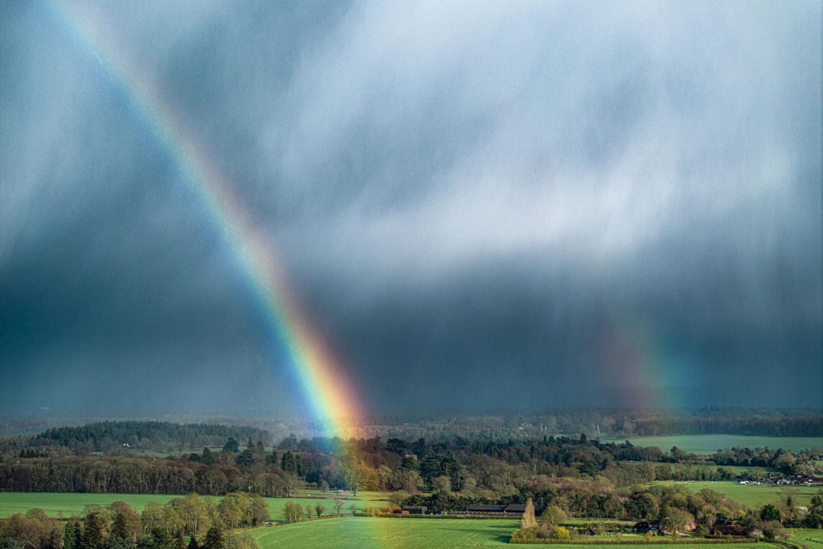 A vivid primary rainbow descends through the base of a storm cell above the Thames Valley near Henley, its foot illuminated by a shaft of low evening light breaking beneath the advancing cloud. The aerial vantage compresses the arc against a dark curtain of rain, while spring fields and deciduous woodland spread beneath in fresh growth. The convergence of storm shadow and spectral colour gives the image an uncommon intensity.