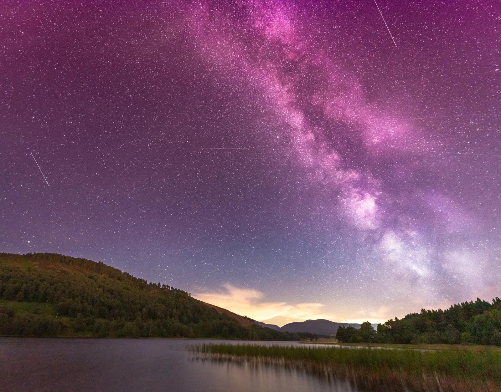 The Milky Way core rises above a wooded Highland loch on a clear August night, its magenta-violet galactic band arching from horizon to zenith. A warm afterglow lingers behind the distant Cairngorm plateau while multiple satellite or meteor trails streak the deep-sky field. Still water in the foreground mirrors the darkness of the scene without resolving a clean reflection.