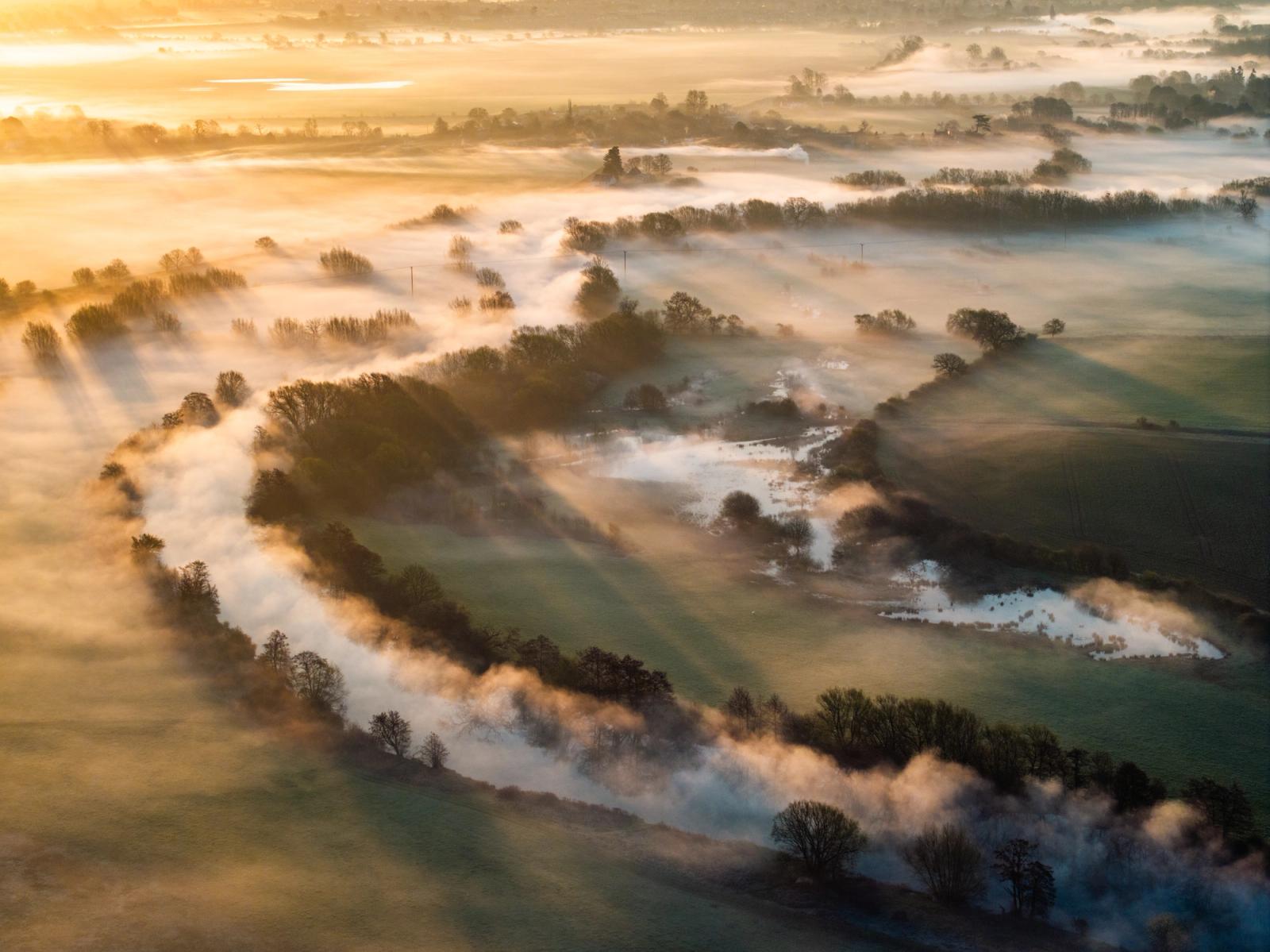 The River Thames is submerged beneath a temperature inversion at dawn, the mist pooling along the watercourse and tracing every curve of the floodplain. Bare riparian trees emerge as silhouettes through the vapour while crepuscular rays fan across the scene from low in the west. Flooded fields catch the light between the fog banks, giving the aerial view an almost topographic clarity.