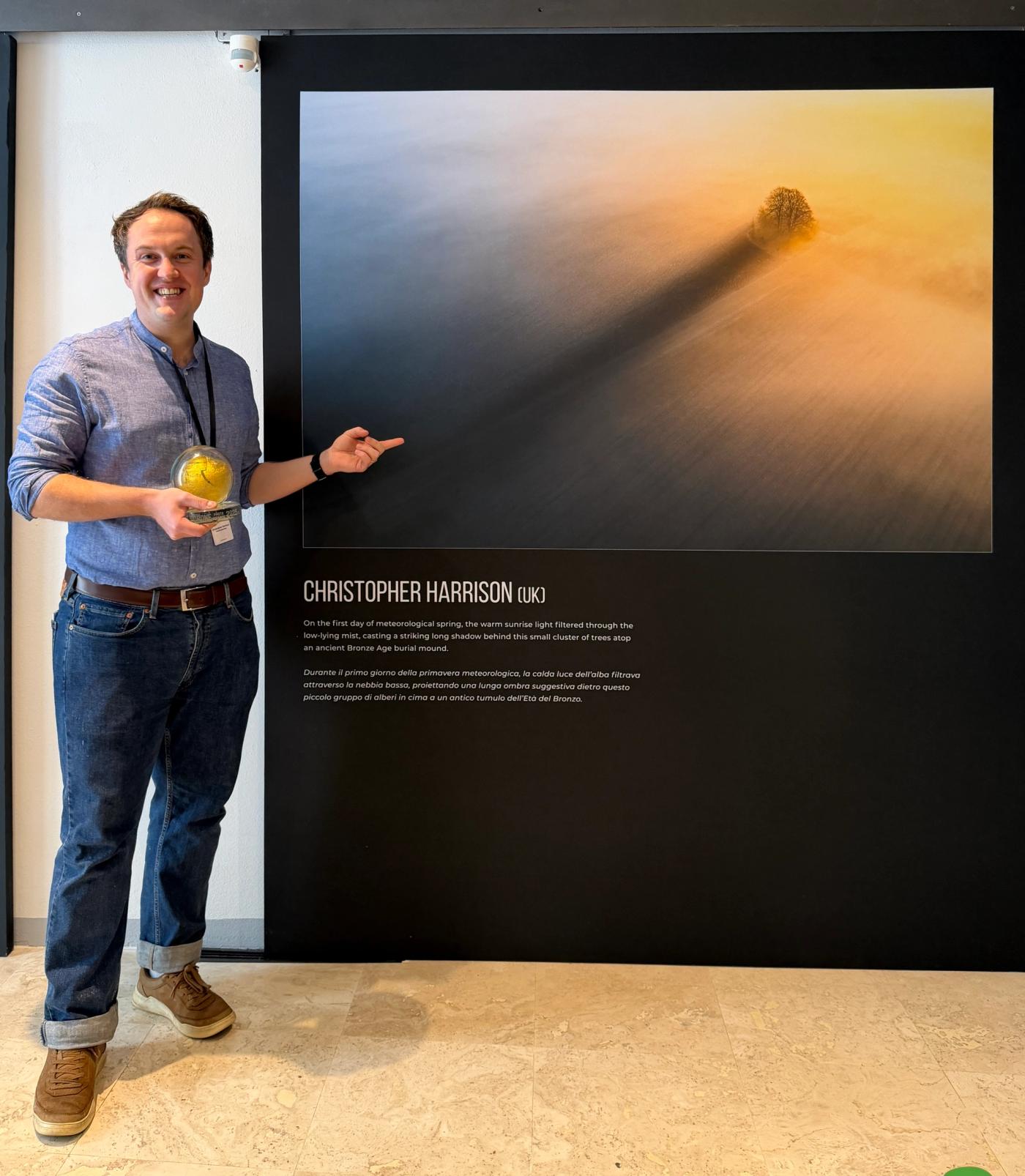 Christopher Harrison with his gold 1st prize trophy beside his exhibited photograph The Long Shadow in the Siena Natural History Museum after winning the Nature category of the Drone Photo Awards.