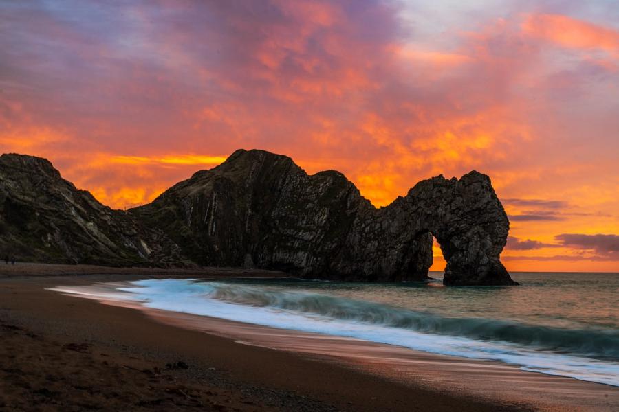 Durdle Door is silhouetted against a vivid winter afterglow on the Jurassic Coast, Dorset. A moderate long exposure softens the incoming waves without erasing their form, preserving the diagonal sweep of the shoreline. The arch frames a sliver of lit horizon, the only gap in the darkening headland mass.