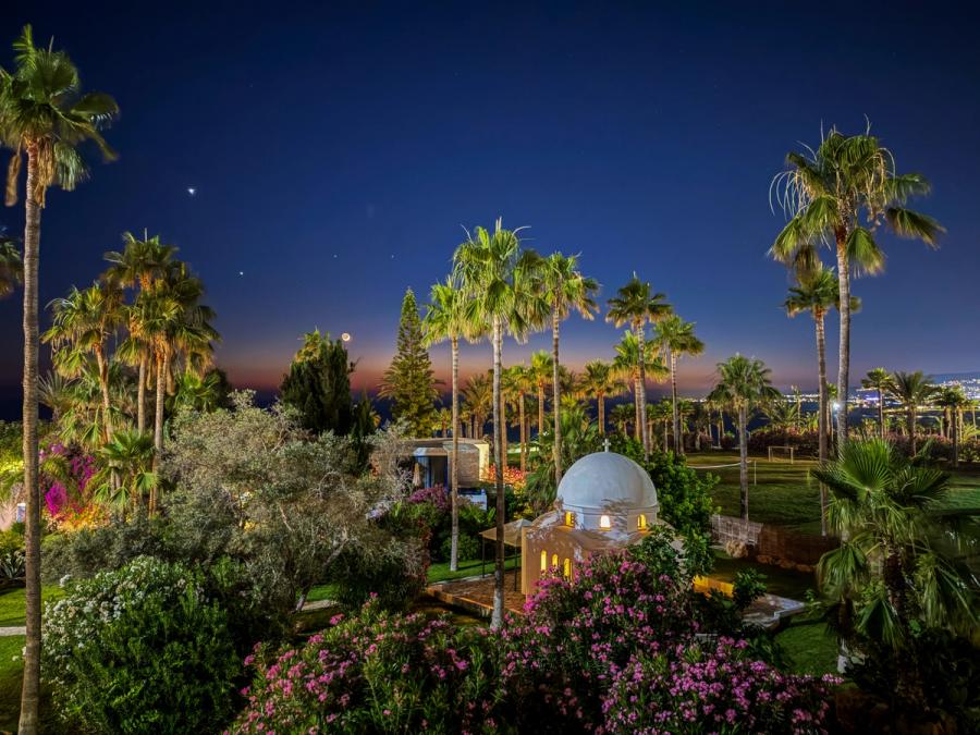 A whitewashed chapel dome sits among manicured gardens at blue hour near Chlorakas, Cyprus. A crescent moon and two planets hang in the deepening sky above the horizon glow, while artificial lighting picks out bougainvillea and palm canopies below. The scene reads more as resort documentation than composed landscape work.