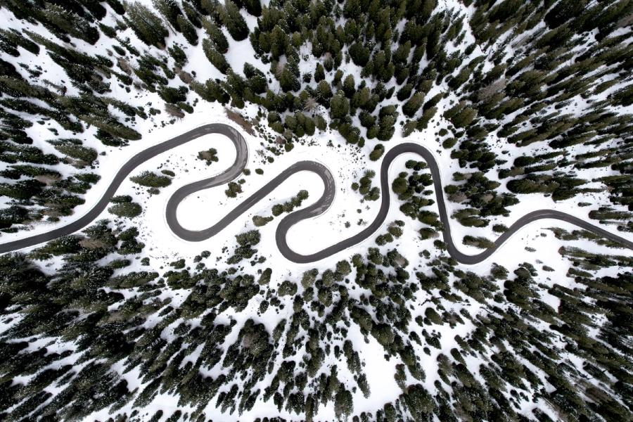 A hairpin mountain road traces repeated switchbacks across a snow-covered alpine slope near Cortina d'Ampezzo, the dark asphalt cutting a rhythmic sinuous line through conifer forest. Shot directly overhead at midday, the nadir perspective strips away horizon and depth, reducing the scene to graphic pattern. The tonal contrast between cleared road, unbroken snow, and dark evergreen canopy gives the composition a near-typographic quality.