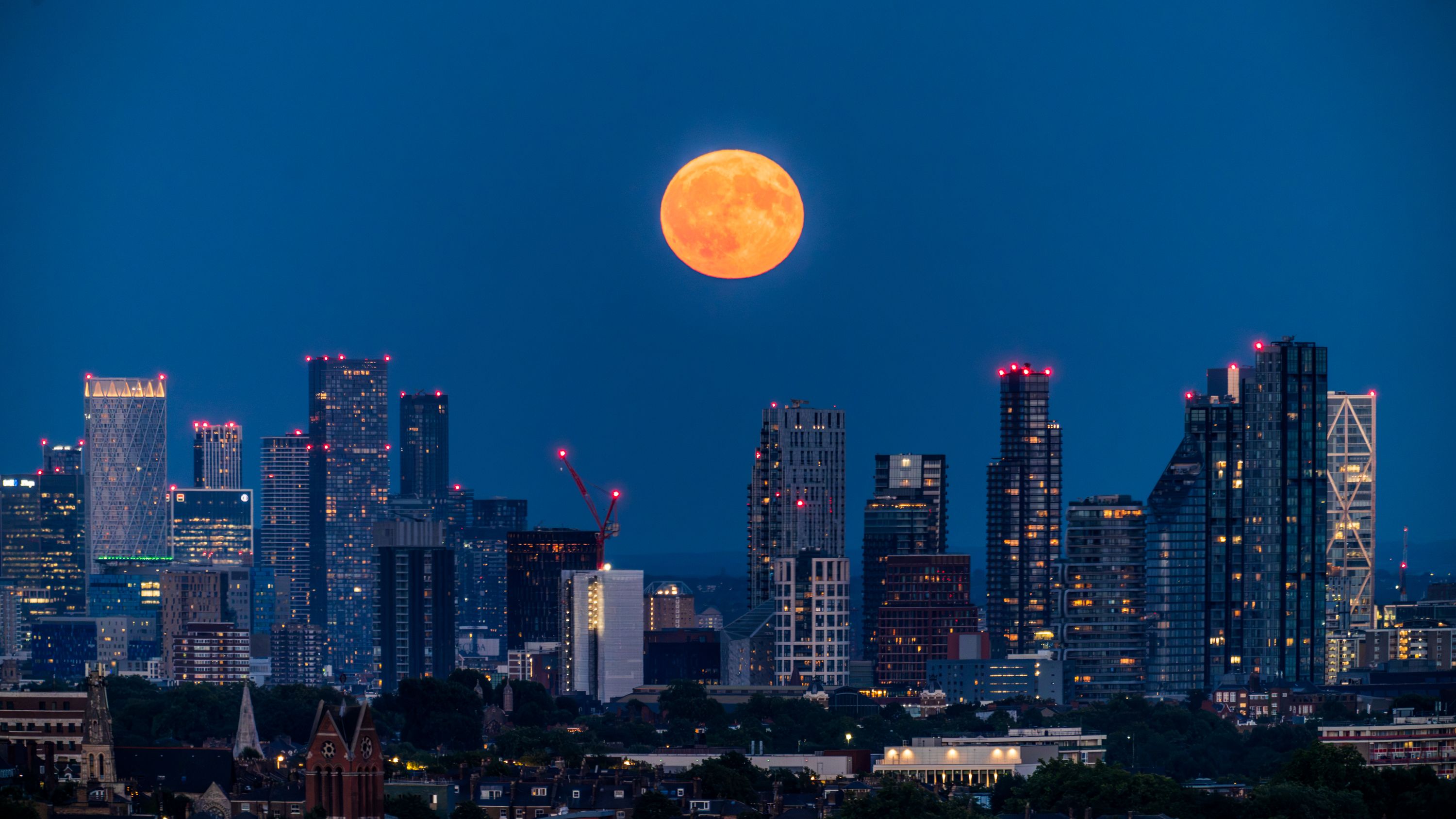 The full moon rises over the growing and glowing London skyline of Canary Wharf during blue hour after sunset. 