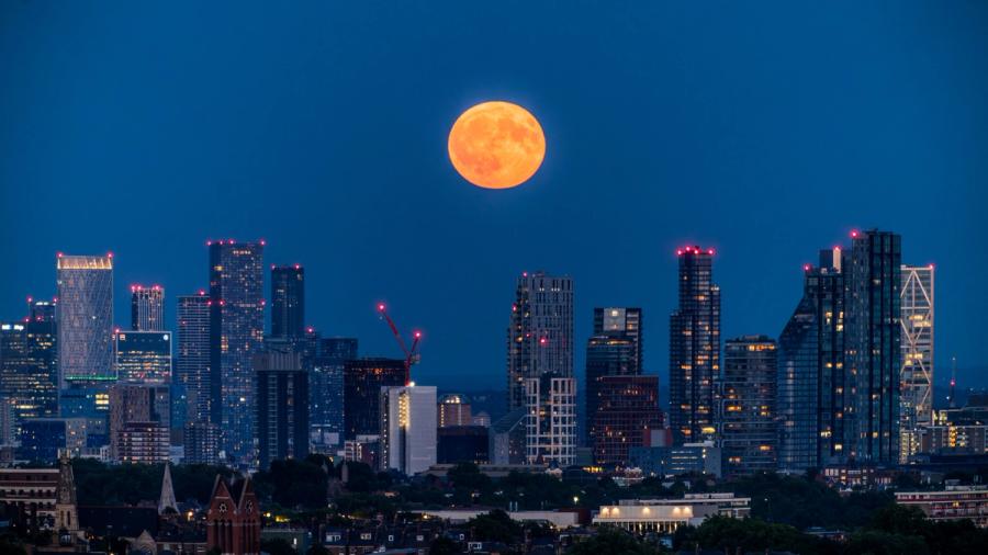 The full moon rises over the growing and glowing London skyline of Canary Wharf during blue hour after sunset. 