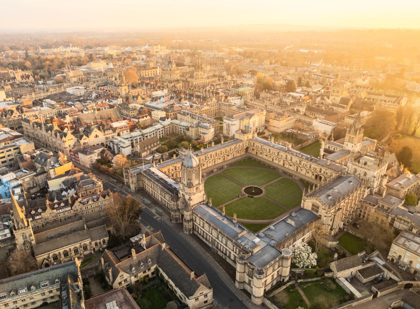 Christ Church College seen from altitude at late-afternoon golden hour in early spring, its quadrangle lawn and central fountain sharply resolved against the honey-coloured limestone of Oxford's medieval core. The warm haze softens the city's edges without obscuring the collegiate geometry that defines the foreground.