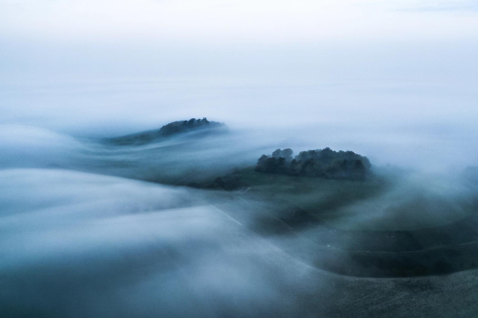 A temperature inversion fills the valleys around Brightwell Barrow and the Wittenham Clumps in Oxfordshire with rolling fog at dawn, leaving two wooded hilltops as dark islands adrift in a monochrome sea. The long-exposure drone frame records the fog's motion as fluid, directional streaks, compressing sky and ground into a single tonal plane. Captured in mid-October near Abingdon-on-Thames, the image sits at the threshold between landscape and pure abstraction.