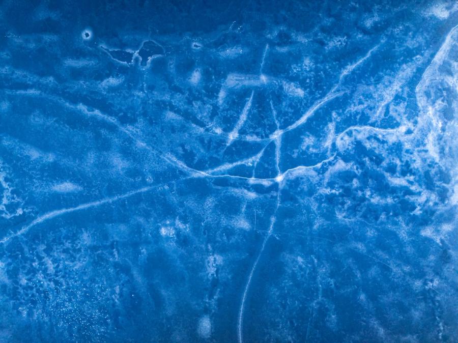 A network of stress fractures radiates across the surface of frozen ground near Nethy Bridge, Cairngorms, captured from directly above in late December. The monochromatic blue palette â deepened by low winter light and the drone's angle of incidence â renders the ice as something closer to celestial cartography than familiar landscape. Frost crystals and air pockets beneath the surface add granular texture at the margins.