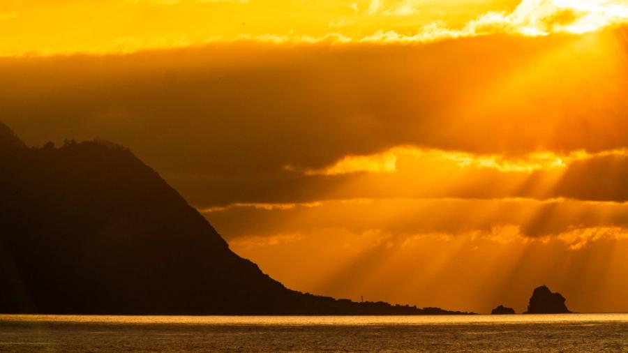 Crepuscular rays break through storm cloud above a volcanic headland, casting the sea in deep amber. Rock stacks punctuate the mid-distance, their silhouettes anchoring the composition against the luminous sky. The light is fleeting and the tonal range extreme, caught in the last minutes before sunset.