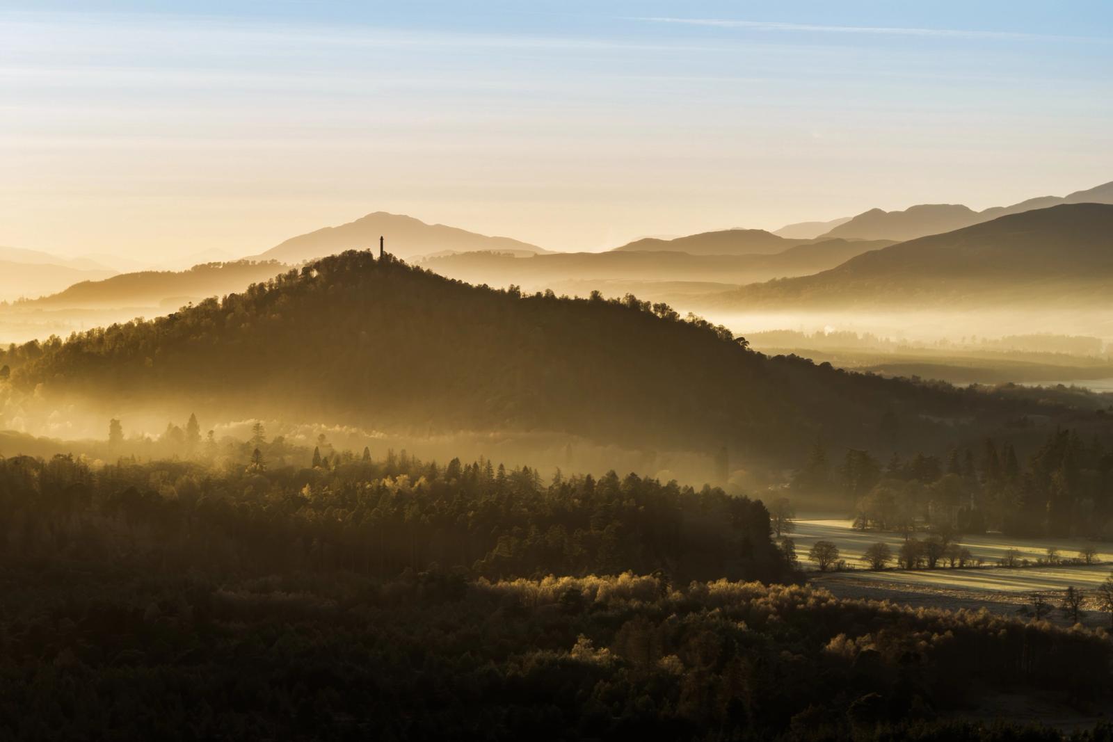 A temperature inversion pools valley mist between forested ridges near Aviemore, the Cairngorms, on a December morning. A solitary monument crowns the nearest wooded hill, its silhouette the only vertical interruption in a composition built entirely from receding planes of light and atmosphere. The distant Highland peaks dissolve into a warm, pale sky, compressing depth into something almost painterly.