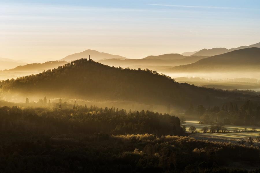A temperature inversion pools valley mist between forested ridges near Aviemore, the Cairngorms, on a December morning. A solitary monument crowns the nearest wooded hill, its silhouette the only vertical interruption in a composition built entirely from receding planes of light and atmosphere. The distant Highland peaks dissolve into a warm, pale sky, compressing depth into something almost painterly.