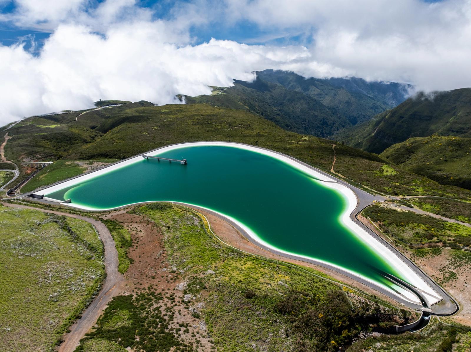 An elevated reservoir on the Paul da Serra plateau, Madeira, its pale concrete liner forming a sinuous white border against the deep teal water. Low cloud clings to the mountain ridge beyond, softening the transition between the engineered basin and the laurisilva-covered slopes. The organic curve of the structure from altitude reads almost as a natural form pressed into the upland terrain.