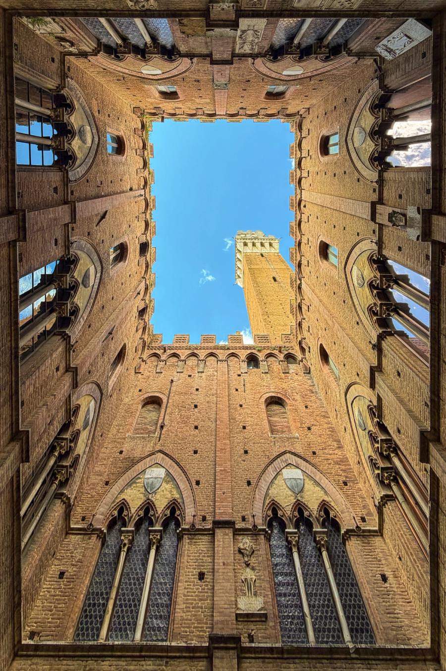 Looking straight up from the inner courtyard of the Palazzo Pubblico, Siena, the Torre del Mangia rises through a rectangular opening of medieval brickwork into a clear September sky. The symmetry of the Gothic arcade below anchors the composition, compressing centuries of civic architecture into a single vertical frame.