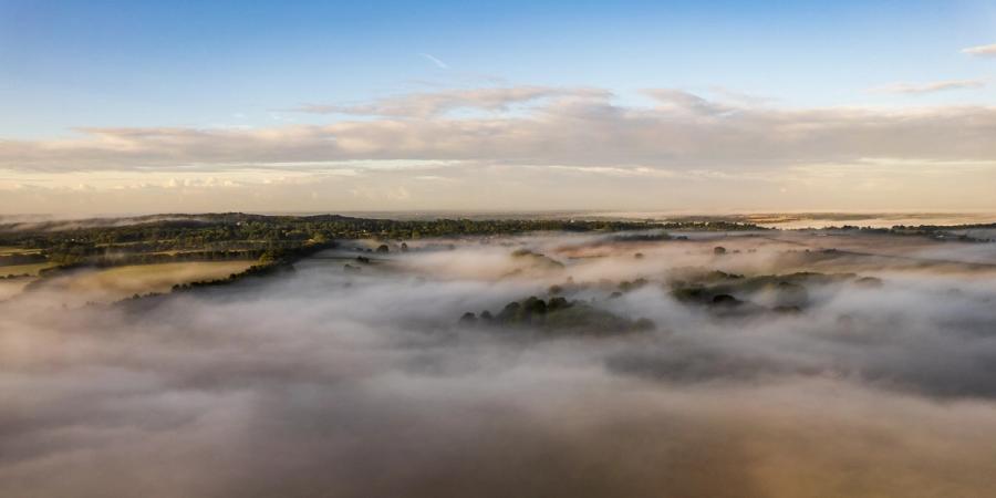 A temperature inversion pins a shallow fog across the Thames Valley lowlands near Maidenhead, submerging the fields while tree canopies and distant wooded ridges remain above the surface. Captured at first light in early September, the aerial vantage reveals the fog as a continuous, almost liquid plane, broken only by the crowns of mature broadleaf trees. The soft, diffuse illumination holds the scene in a quiet, pre-colour state.