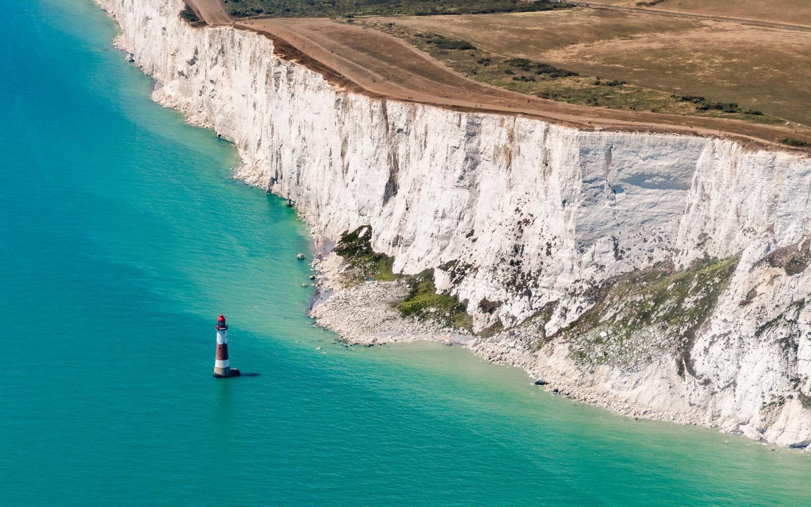 Beachy Head lighthouse stands isolated in calm English Channel waters below the sheer white chalk face of the Seven Sisters cliffs, East Sussex. The aerial perspective flattens the geometry of cliff, shore, and sea into near-abstract bands of colour, the lighthouse reduced to a small vertical accent against the turquoise. Midday light removes shadow relief from the cliff face, flattening texture in the brighter sections.
