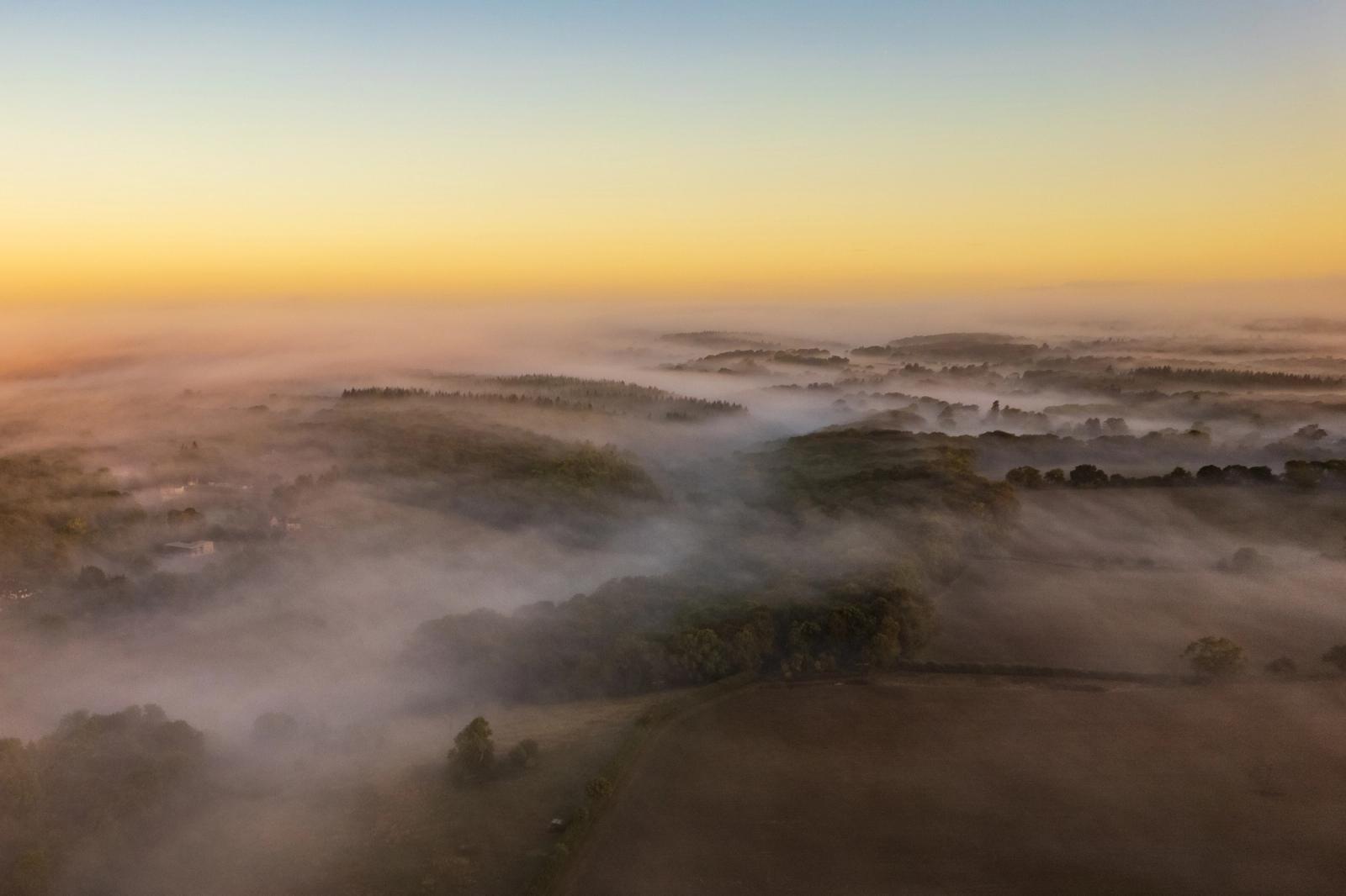 Ground mist pools in the valleys and hedgerow corridors of the Chiltern escarpment south of Henley-on-Thames, photographed from altitude at first light in early September. Tree canopies emerge as islands above the fog, their forms softened by the warm pre-sunrise glow. The layered recession of mist and ridge creates a quiet sense of depth across the rolling farmland.