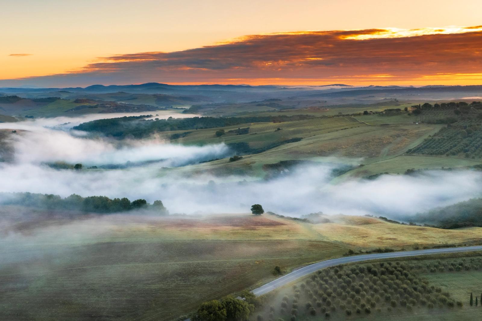 Ground-level inversions pool between the rolling hills of the Val d'Orcia at dawn, the mist threading between tree lines and farmland as the sky ignites above the distant Apennine ridge. Shot from altitude, the layered recession of fog, hill and cloud gives the scene its depth. Autumn colour is still emerging in the canopy below.