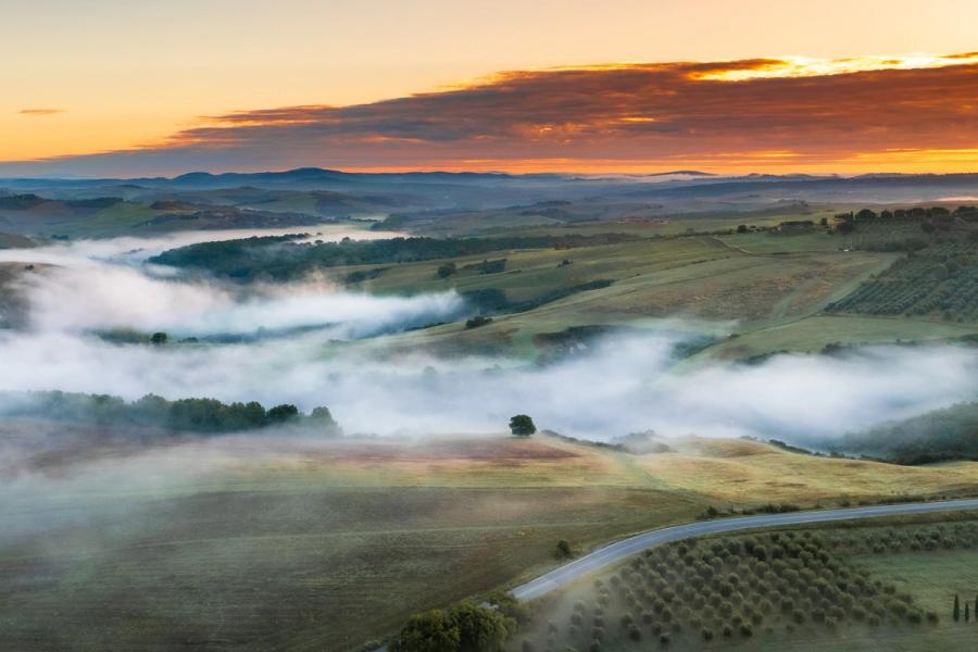 Ground-level inversions pool between the rolling hills of the Val d'Orcia at dawn, the mist threading between tree lines and farmland as the sky ignites above the distant Apennine ridge. Shot from altitude, the layered recession of fog, hill and cloud gives the scene its depth. Autumn colour is still emerging in the canopy below.