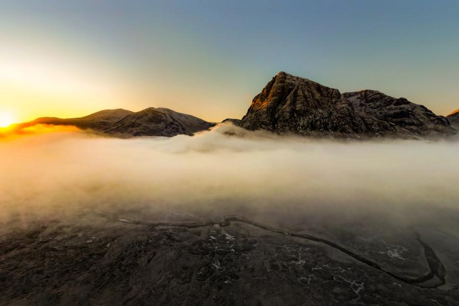 A temperature inversion fills the glen floor with dense fog at sunrise, leaving two rocky summits isolated above the cloud. The winding road beneath the mist surface anchors the composition, hinting at scale and the valley's geometry. Shot in early winter, the frost-dusted ridgelines catch the first direct light while the foreground terrain remains in cold shadow.