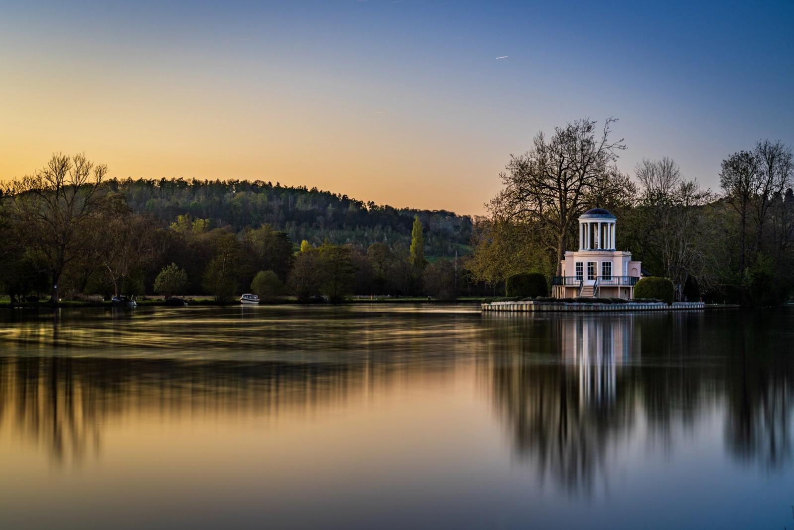 A neoclassical riverside temple (Temple Island, Henley-on-Thames) catches the last warmth of an April sunset, its columns and dome reflected in long-exposure-smoothed water. The gradient from amber horizon to deep blue zenith frames the building with quiet formality. A vapour trail and moored boats add understated human presence without disrupting the stillness.
