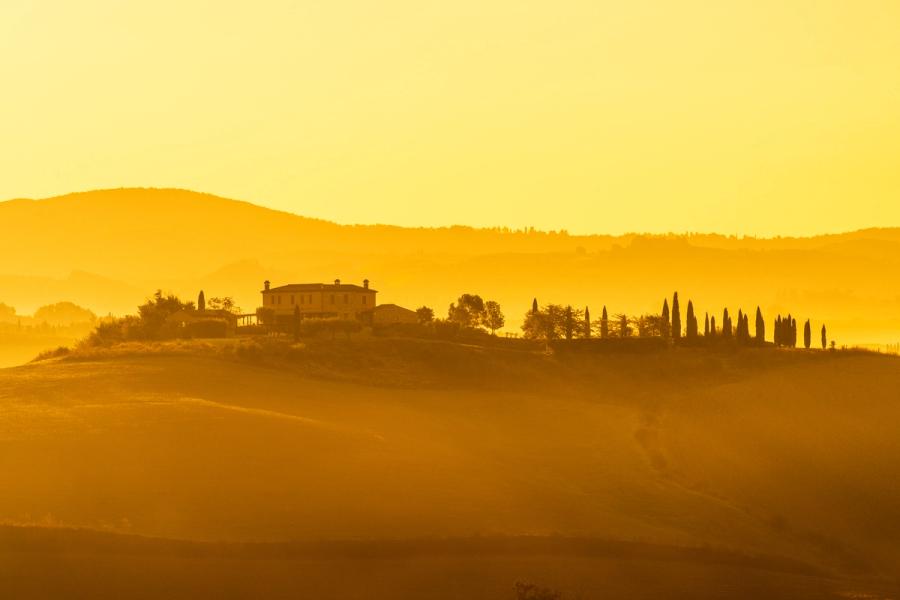 A Tuscan farmhouse and a row of Italian cypress trees sit on a rolling hillside, rendered almost entirely in silhouette against a dense amber haze. Layered bands of mist separate the undulating foreground from a receding ridge line, compressing the scene into a series of warm, near-monochromatic planes. The image is unmistakably Mediterranean in character, distant from the photographer's usual British subject matter.