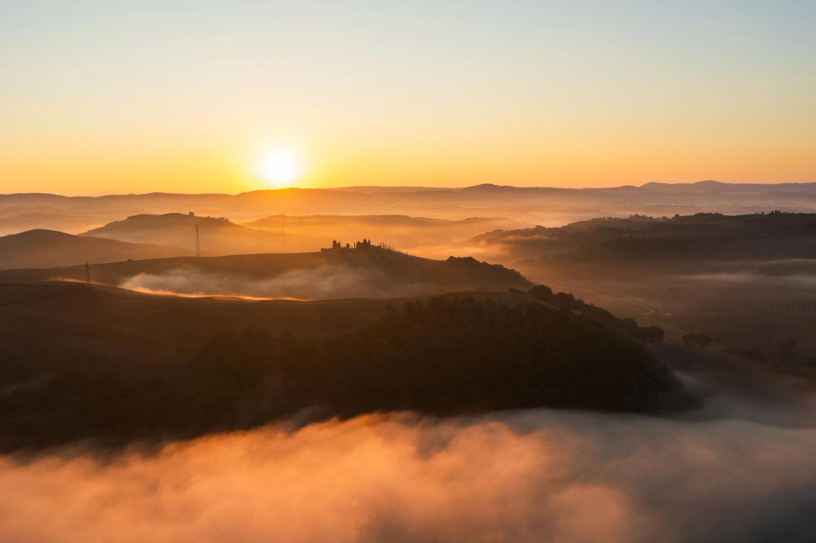 A temperature inversion fills the Val d'Orcia valleys with layered mist at dawn, isolating a hilltop farmstead and its cypress avenue as a lone silhouette against the rising sun. Aerial perspective compresses the rolling Sienese hills into receding tonal bands, the foreground fog mass anchoring the frame. Captured in the last minutes before the inversion burned clear.