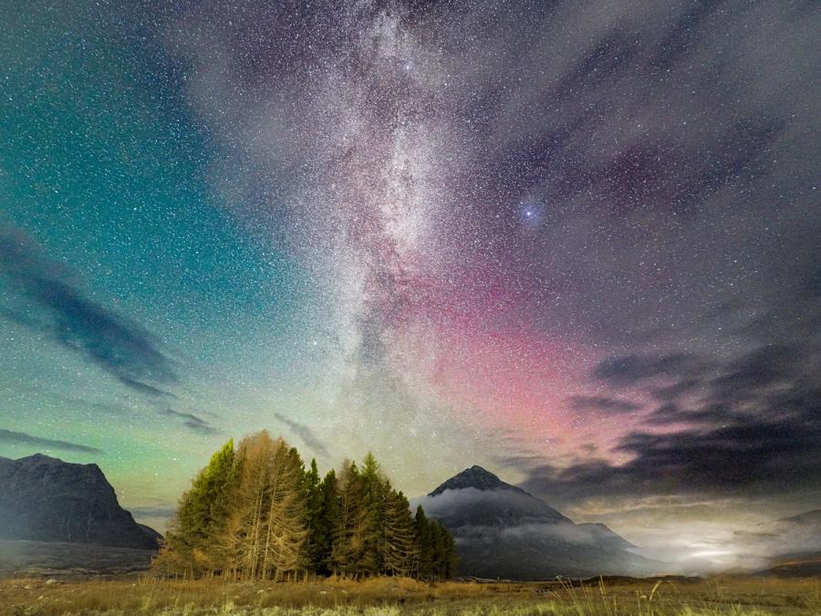 The Milky Way rises vertically above a stand of conifers in a Scottish glen, its core flanked by faint auroral red on the right horizon. Low cloud pools around the base of a pyramidal peak, while green airglow tints the left sky. The coincidence of galactic core, aurora, and valley mist in a single frame is rare and well-judged.