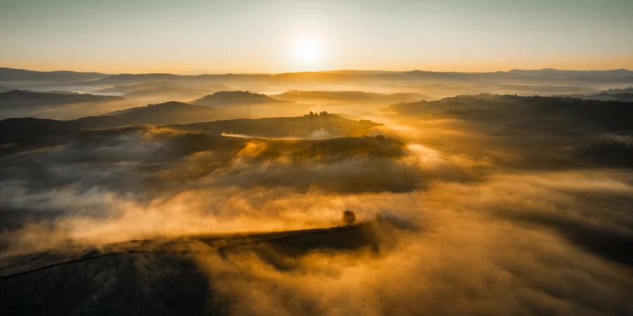 Low-lying fog pools between the rolling hills of the Val d'Orcia landscape at dawn, lit from behind by a rising sun still close to the horizon. A lone farmstead on an isolated hillock breaks the surface, anchoring the composition amid the dissolving mist. The image was captured from altitude, compressing the layered landforms into a quiet, painterly sequence of receding planes.